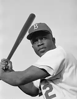 A black and white portrait photo of Jackie Robinson from the waist up. He has a dark complexion and is wearing a white baseball jersey and baseball cap with a stylized letter "B" on it. He is holding a baseball bat and posed to swing.