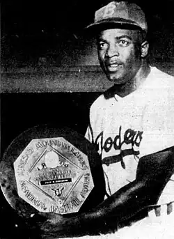 A black man in a Brooklyn Dodgers uniform and a cap with the letter "B", smiling while holding a plaque.