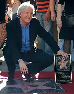 James Cameron squats by his new star, left hand propping up his plaque