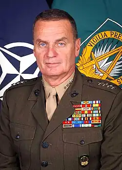 Full face color portrait photo of General James L Jones - a caucasian male with short dark hair and blue eyes; wearing US Marine Corps service A uniform - khaki shirt with four silver stars on the collar and khaki tie; wearing dark green jacket with 4 silvers stars on each shoulder epaulet, dark eagel clog and anchor emblem on both lapels, and multiple colorful military ribbons on the left chest.