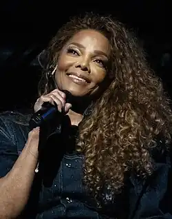 Janet Jackson, on stage, with big curly hair looking to her right