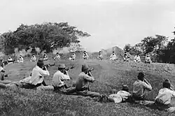 A dozen prisoners sit bound at a distance as Japanese soldiers in the foreground kill them