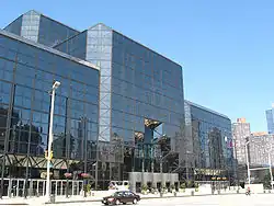 The Javits Center, a building of dark tinted glass, stands over a city street. The corners of the building are smoothed at 45-degree angles.