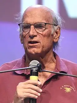 Head shot of Ventura speaking into a microphone, wearing glasses and a red shirt