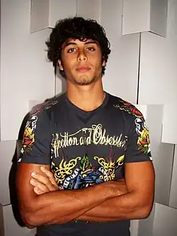 A man with curly dark hair and light stubble stands with his arms crossed, wearing a short-sleeved graphic T-shirt against a white geometric background.