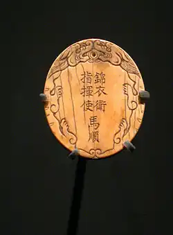 An oval, light-colored plaque (possibly made of bone or ivory) featuring Chinese characters carved in two vertical columns: "Embroidered Uniform Guard Commander Ma Shun". The inscription is framed by stylized carvings of clouds or dragons at the top and sides.