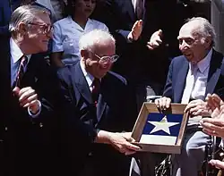 Johnny Grant, wearing glasses and a dark blue suit, and Joe Pasternak, also wearing a dark blue suit, hold a Walk of Fame award that features a replica star as a crowd around them clap and look on