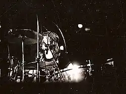 A black and white photograph of John Bonham wearing a headband and behind the cymbals of a drum kit
