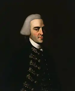 Half-length portrait of a man with a hint of a smile. His features suggests that he is in his 30s, although he wears an off-white wig in the style of an English gentleman that makes him appear older. His dark suit has fancy embroidery.