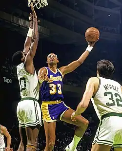 A bald black person, wearing a gray suit and a tie, looks to the left.