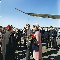 Kennedy and the First Lady, dressed in a pink outfit and holding a bouquet, depart from Air Force One and greet the welcomers