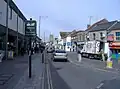 Street scene showing shops on left and right, with cars and vans on road. On the left hand pavement is a sign saying welcome to Keynsham high street.