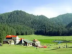 Daytime sunlit view of a verdant bowl-shaped meadow with a small pond in a valley rimmed by several prominent hills covered in evergreen forest. At centre-left and looking out on the meadow, a small cluster of tourist lodges with pitched roofs, one a bright red. Around them and the pond are small, gaily coloured gazebos and shade-giving umbrellas. Several dozen tourists and sheep mill about the pond and in the meadow.
