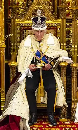 A colour photograph of Charles III during his coronation, showing him on a holden chair wearing his coronation robes and the Imperial State Crown, while also holding some document in his hands.