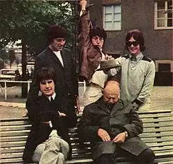 On a bench in a park sit five men, two seated and three standing behind the bench. Clockwise from left is a man in a black suit with khaki pants; a man in a black suit with black pants; a man wearing a brown coat with khaki pants and raising his arm jokingly as if to stab the sleeping man below him; and a man wearing a grey/light green coat and khaki pants and holding out a hat above the sleeping man's head. The sleeping man is wearing green.