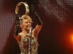 An image of Kylie Minogue, with one of her raising hands holding a tambourine. The microphone in a microphone stand is seen infront of Minogue.