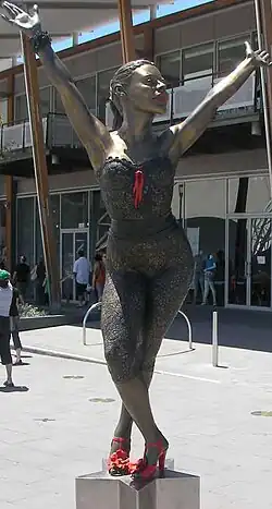 A bronze statue resembling Kylie Minogue, on a star-shaped pedestal, portrays her in a dancing pose. Her legs are crossed, and she bends at the waist, with both arms stretched above her head. The statue stands in a public square in front of a modern glass building, and several people are walking.