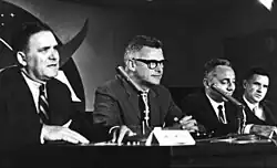 A black and white photograph of four men seated at a table with a mike in front of them