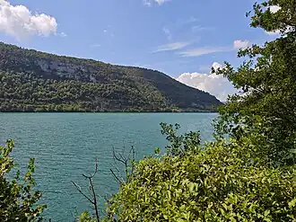 A turquoise lake bordered by forested slopes and limestone cliffs in the Jura Mountains of France.