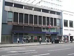 A grey building with blue panels and a dark blue, rectangular sign reading "LANCASTER GATE STATION" in white letters and people in front