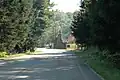 A road through a forest leads to a fieldstone building with a chmney and steep roof next to a stop sign