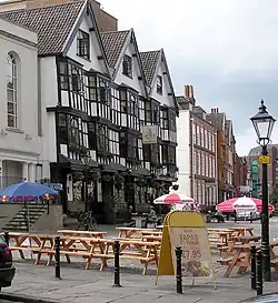A seventeenth-century timber-framed building with three gables and a traditional inn sign showing a picture of a sailing barge. Some drinkers sit at benches outside on a cobbled street. Other old buildings are further down the street, and in the background, part of a modern office building can be seen.