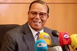 An elder black gentleman, with short cropped black hair and wearing glasses, sitting before a podium with microphones in front of him.
