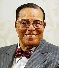 An elder black man with short cropped black hair, wearing glasses, looking out towards the viewer. He is wearing a white shirt, grey suit jacket, and red bowtie.