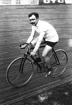A black-and-white photograph of a man with three-colored sweater and shorts with a moustache sitting on a bicycle.