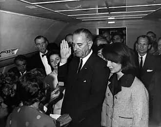 Lyndon B. Johnson raises his hand above an outstretched Bible as he is sworn in as President as Air Force One prepares to depart Love Field in Dallas. Jacqueline Kennedy, still in her blood-spattered clothes (not visible), looks on.