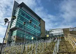 A glass and steel building on the left of a photograph, as viewed from the bottom of a large set of outdoor stairs leading up to it. The view of the building is partially obscured by lamp posts and the stairs are split up by steel railings