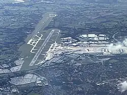 An aerial view of a large airport; from left to right there are two parallel runways and then a series of terminal buildings around a central flat area. In the foreground the airport backs onto urban sprawl but in the background it meets fields.