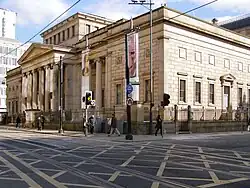 A neopalladian cuboid building as viewed centred on its corner from across the street. Halfway down the facade is a portico, and there is a central cuboid atrium that rises in the centre of the roof above the main roof height. There are regularly arranged sash windows in two stories.