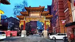 A Chinese arch structure on a street with a brick building to the right and no building to the left. There is a tree in the background and it is a clear early evening sky.