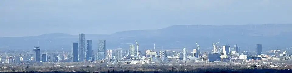 A skyline of a city against a blue sky and tall hills in the background. There are around nine skyscrapers distributed fairly evenly across the skyline with many cranes also visible. In the foreground there is a river with trees on both banks.