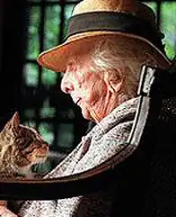 A color photograph of Marjory Stoneman Douglas late in her life. She is shown in profile, seated, with a cat on her lap. She is white-haired, tanned, and wrinkled. She wears a lapelled jacket and low-brimmed straw hat. She and the cat gaze at each other lovingly.