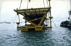 A partial section of a wooden ship lying inside a massive steel frame is suspended just about the water with a partial view of a barge on the left and small boats in the background.