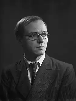 A black-and-white photo of an elderly, balding man with swept-back hair, wearing a suit; he faces the camera.