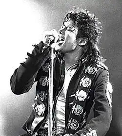 Black and white picture of a man with black hair in a ponytail singing to a microphone; he's wearing a black jacket with buckles.