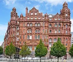 A large red-brick building with curved windows as viewed from a short distance away. There are green trees in front and a blue sky behind. The building curves round on both sides to follow the road.