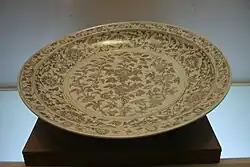 A large, shallow porcelain dish resting on a dark wooden base, viewed from an angle. The dish has a light body decorated with intricate floral and scrollwork patterns rendered in a dense, monochromatic underglaze brown or sepia pigment.