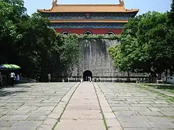 A wide stone path leads towards a tall grey brick wall with a central arched entrance, surmounted by a red building with a double-tiered yellow roof, framed by trees on both sides.