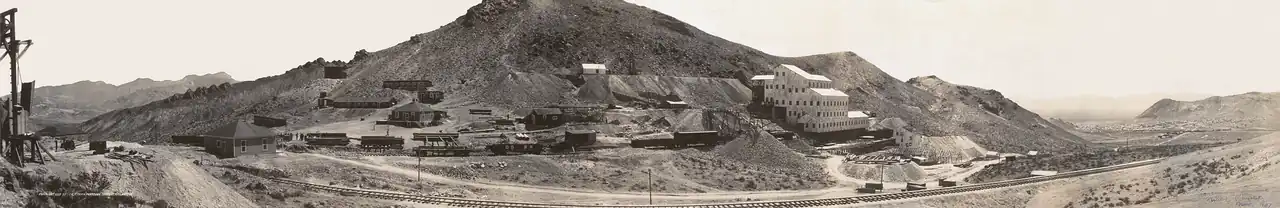 A large mill, five or six stories high and built on the side of a mountain, is surrounded by mine tailings, rail cars, piles of lumber, and a variety of smaller buildings. A rail line leads from the mining operation toward a city nestled between hills in the distance.