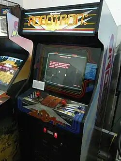 A vertical wooden structure with a visual display unit embedded in the front side. The sides have white panels with blue and red artwork that reads 2084, and the top front panel is a backlit marque that reads: "Williams. Robotron: 2084". Two red joysticks with a pair of blue buttons to the right are below the monitor, which displays a pixelated game screen.