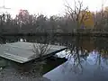 A wooden dock on the shore of a small lake in which autumnal trees are reflected