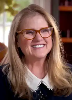 A middle-age woman with long, blonde hair and glasses looking at a camera and smiling