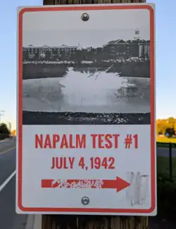 Photo shows a metal sign bolted to a wooden telephone pole with a road, field, and trees in the background. The sign reads "Napalm test #1 / July 4, 1942" with an arrow pointing to the field to the right. Above the text is a black and white photo of an explosion in the field, with the buildings of Harvard University seen in the background.