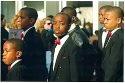 A group of black male adolescents, with shaven heads, wearing black suit jackets, white shirts, and red bowties.