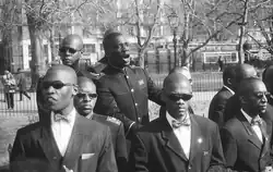 A group of young black men, wearing suits and with shaven heads, standing outdoors. One is in the centre, speaking aloud. The others surround him, facing outward. Although the speaker does not wear sunglasses, the others all do.