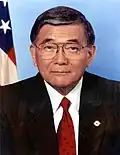 Older Japanese American man with glasses wearing a suit with a red tie with the US flag behind him